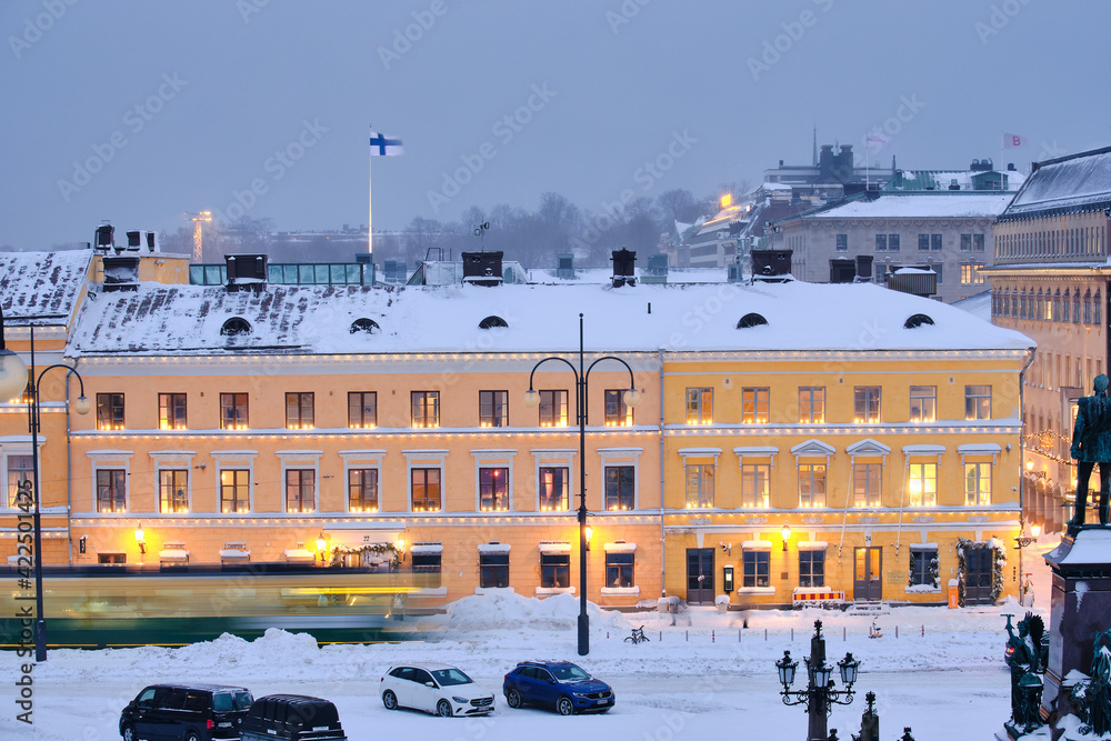 Helsinki, Finland - January 13, 2021: The tram is moving on the Senate Square during the strong snowstorm. The blue building is Helsinki City Museum