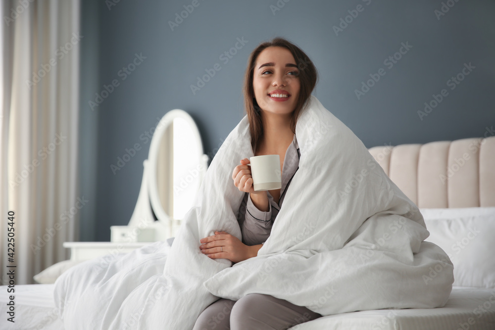 Beautiful young woman wrapped with soft blanket holding cup of hot drink on bed at home Beautiful young woman wrapped with soft blanket holding cup of hot drink on bed at home