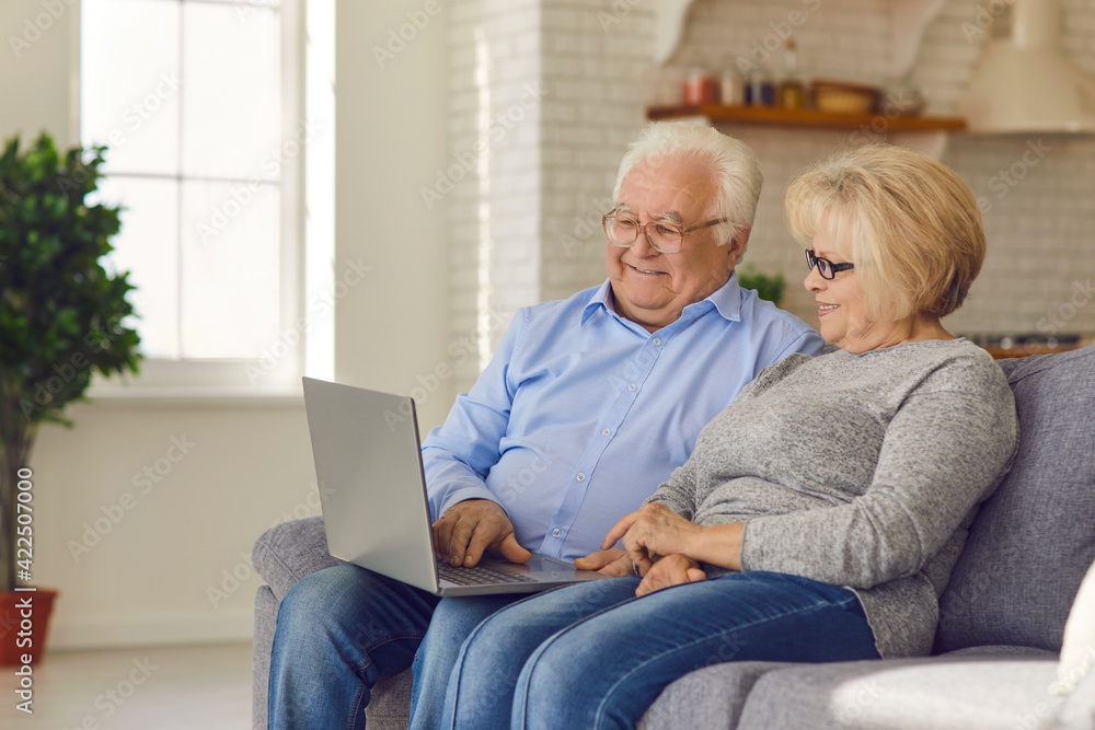 Laughing senior happy couple sitting on sofa at home and watching funny movie or online show on laptop together. Elderly people happy lifestyle and modern technologies concept