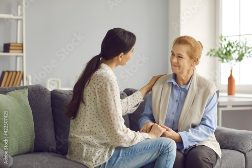 Happy senior mother and young grown-up daughter sitting on sofa at home, holding hands, talking and looking at each other. Family values, helping elderly relative, love, support, mutual understanding