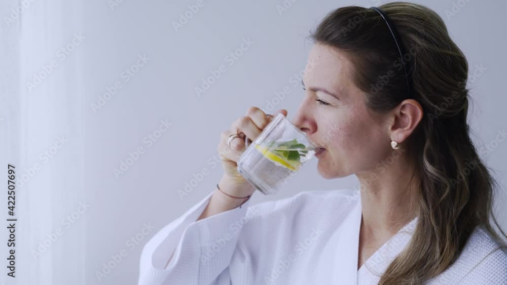 Cheerful long haired woman in white bathrobe drinks delicious infused beverage with lemon and mint in light hotel room in morning close view Cheerful long haired woman in white bathrobe drinks delicious infused beverage with lemon and mint in light hotel room in morning close view