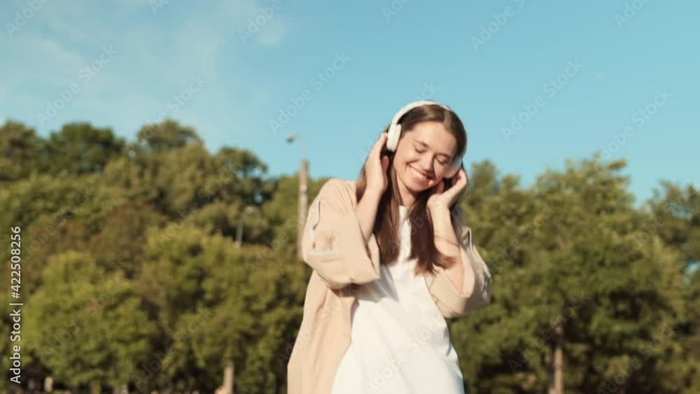 Medium long tracking of young long-haired Caucasian woman wearing white wireless over-ear headphones, smiling, walking and jumping outdoors on sunny summer day Medium long tracking of young long-haired Caucasian woman wearing white wireless over-ear headphones, smiling, walking and jumping outdoors on sunny summer day