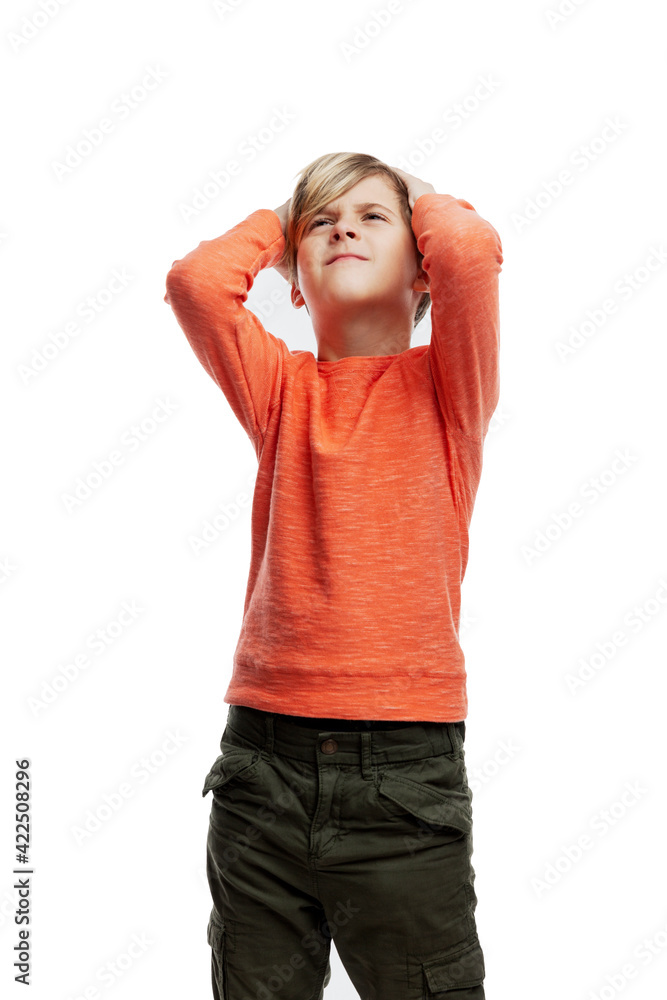 A 9-year-old boy holds his head with his hands and looks up. A guy in an orange sweater with a negative emotion on his face. Isolated on white background. Vertical. A 9-year-old boy holds his head with his hands and looks up. A guy in an orange sweater with a negative emotion on his face. Isolated on white background. Vertical.