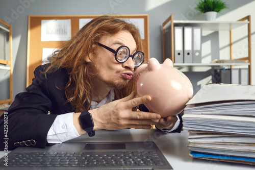 Funny weird looking thrifty man in hilarious round thick lens glasses sitting at office desk with laptop computer and kissing piggy bank. Concept of loving money and saving up photo