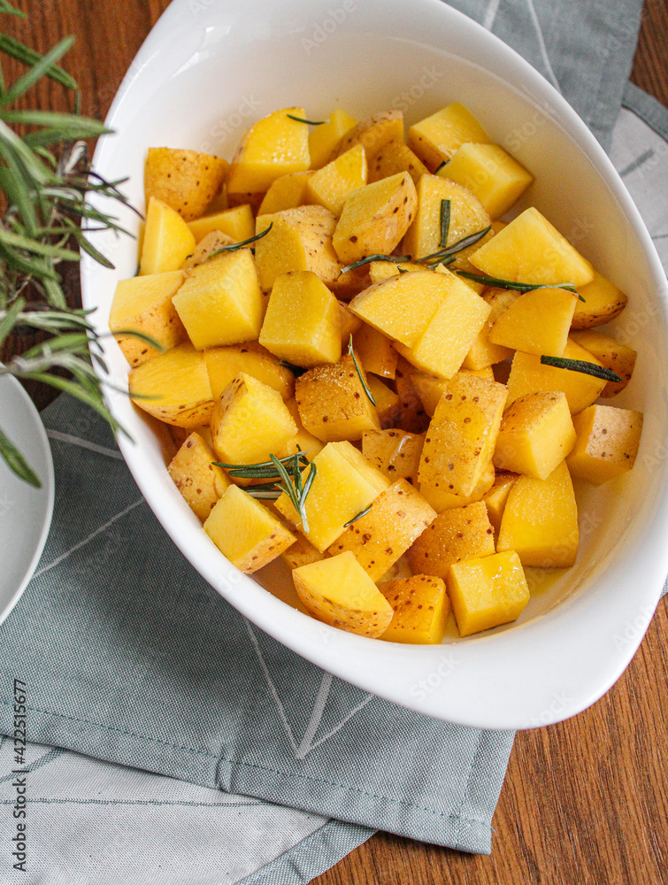 Raw Potatoes ready to be baked with rosemary and olive oil. Preparing roasted potatoes with spices over withe baking dish. Raw Potatoes ready to be baked with rosemary and olive oil. Preparing roasted potatoes with spices over withe baking dish.