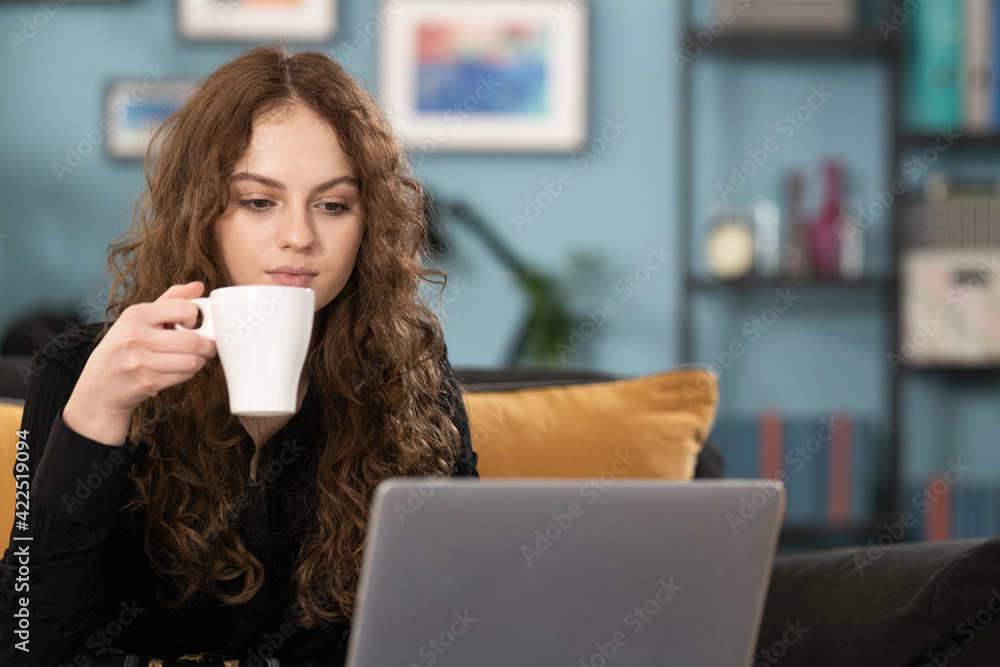 Pretty student girl is studying remotely from home. Female is using laptop browsing websites. The girl is sitting on couch drinking coffee in a cozy living room and looking at the computer screen Pretty student girl is studying remotely from home. Female is using laptop browsing websites. The girl is sitting on couch drinking coffee in a cozy living room and looking at the computer screen