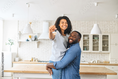 A portrait of hilarious multiracial couple in love in a new property  a husband is holding beautiful wife in his arms  happy couple looks at the camera  a female shows keys of new real estate