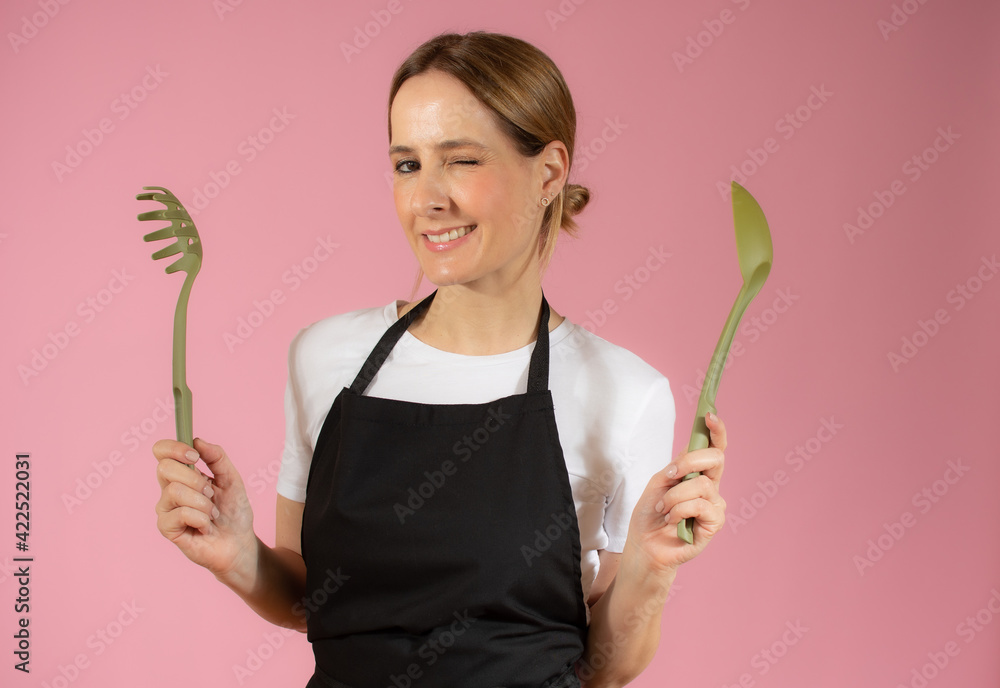 Young caucasian woman holding kitchen utensils against pink background Young caucasian woman holding kitchen utensils against pink background