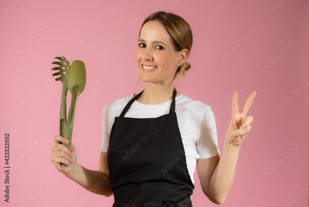 Young caucasian woman holding kitchen utensils against pink background Young caucasian woman holding kitchen utensils against pink background