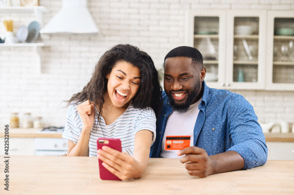 Obraz premium Overjoyed biracial couple shopping together online, excited multiracial woman and man hold smartphone and a credit card, rejoy purchases online, making order, buying something in e-shop