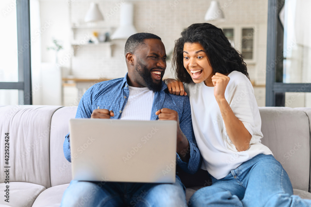 Overjoyed cheerful multiracial couple celebrating good news looking at the laptop screen, young excited woman and man scream happily sitting with computer on the cozy couch at home, won in video game Overjoyed cheerful multiracial couple celebrating good news looking at the laptop screen, young excited woman and man scream happily sitting with computer on the cozy couch at home, won in video game
