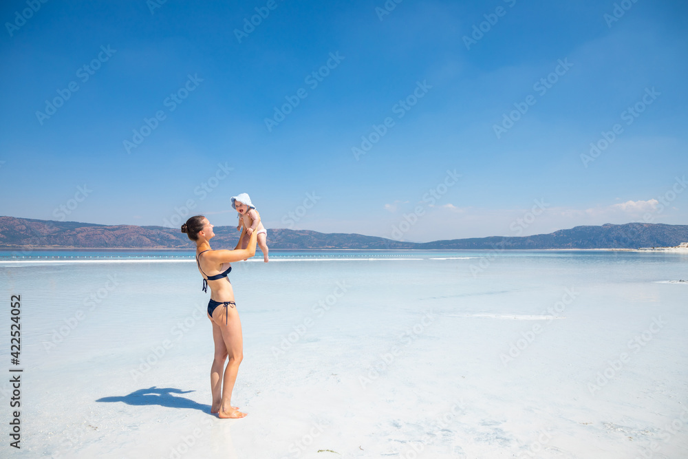 Young woman in swim suit and her daughter little baby girl are having fun on white clay of turquoise crater lake Salda Golu, Turkey