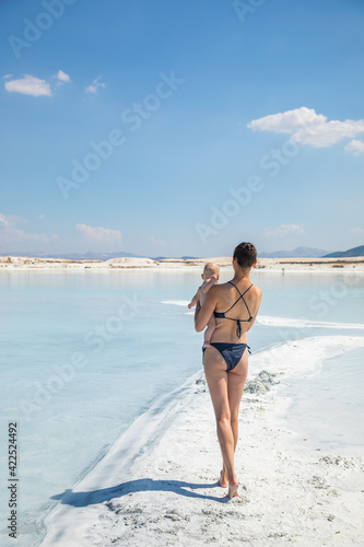 Young woman in swim suit and her daughter little baby girl are walking on white clay of turquoise crater lake Salda Golu, Turkey