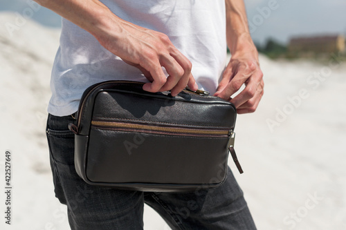 Leather Waist Belt Bag Leather Fanny Pack Men Waist Purse. A young guy in a white T-shirt walks on the sand with a dark brown leather chest bag. Travel concept.