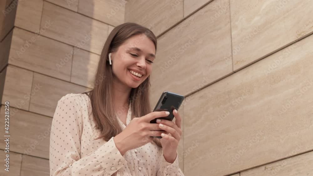 Low angle arc of delighted beautiful Caucasian woman smiling, wearing in-ear wireless headphones, using smartphone by building outdoors Low angle arc of delighted beautiful Caucasian woman smiling, wearing in-ear wireless headphones, using smartphone by building outdoors