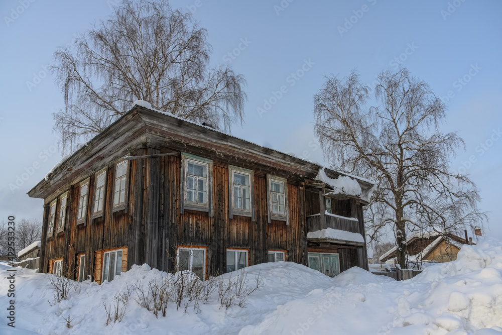 Wooden 2-storey historic house in the provincial town of Cherdyn (Northern Ural, Russia) on a frosty winter day. Traditional urban style of the time, window frames, balconies, tall trees 