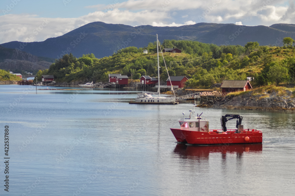 
View from ferry to the small fishermen village with traditional red houses in Helgeland archipelago in the Norwegian sea. Red boat in the foreground