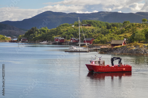  View from ferry to the small fishermen village with traditional red houses in Helgeland archipelago in the Norwegian sea. Red boat in the foreground