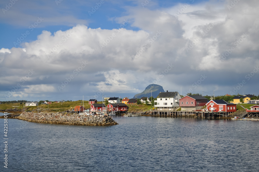 View from ferry to the small fishermen village with red houses on island in Helgeland archipelago in the Norwegian sea on sunny summer morning. Lovund island in background