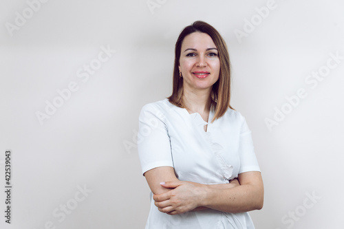 Beautiful young female doctor in a white medical gown, on an isolated white background. The beautician smiles into the camera, arms folded. With place for text or logo.
