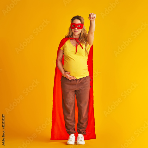 attractive smiling pregnant woman in superhero costume, wearing red mask and cape, stands with her arm outstretched on a yellow background. concept superpowers of a girl, feminism, desire to win