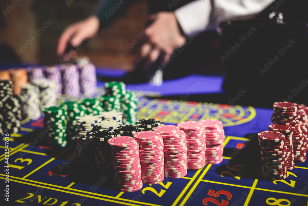 A close-up vibrant image of multicolored casino table with roulette in motion, with casino chips. the hand of croupier, money and a group of gambling rich wealthy people playing in the background A close-up vibrant image of multicolored casino table with roulette in motion, with casino chips. the hand of croupier, money and a group of gambling rich wealthy people playing in the background