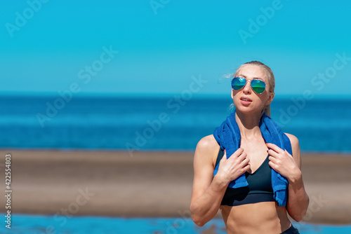 sportswoman with a towel on her shoulders is resting on the beach against the background of the sea after cardio workout. The concept of a healthy lifestyle after a pandemic. 