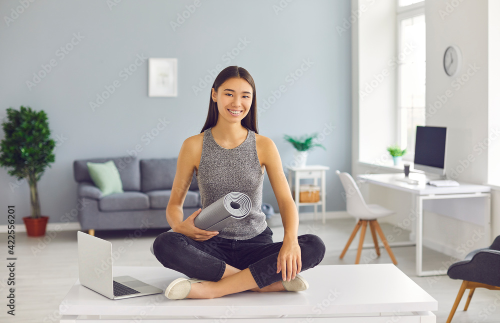 Smiling slender asian girl sitting on the table next to a laptop with a yoga mat in her hands. Smiling slender asian girl sitting on the table next to a laptop with a yoga mat in her hands.