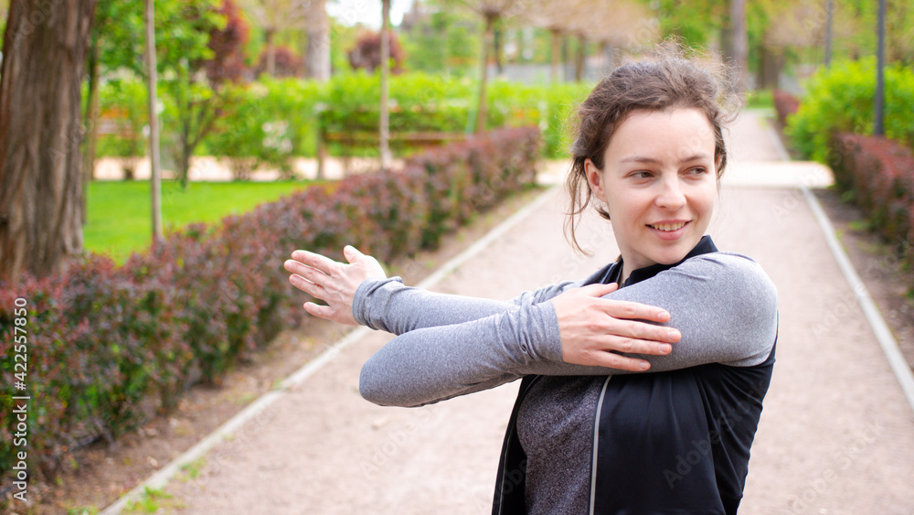 Happy sporty girl jogger stretching arms during morning exercising outdoors before running, standing in urban park in spring day, wearing grey sportwear and looking in distance.