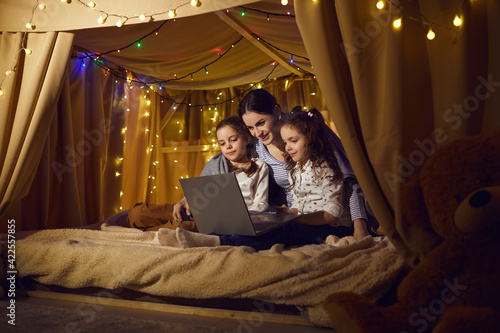 Young caucasian mother with preschool daughters watching movie using laptop online application, making video call, having fun. Parent spending time with children in kids tent at home during evening