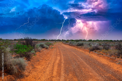 Deserted Australian outback landscape with red dirt road towards horizon with bushes in roadsides and heavy thunderstorm with white purple lightnings on the horizon