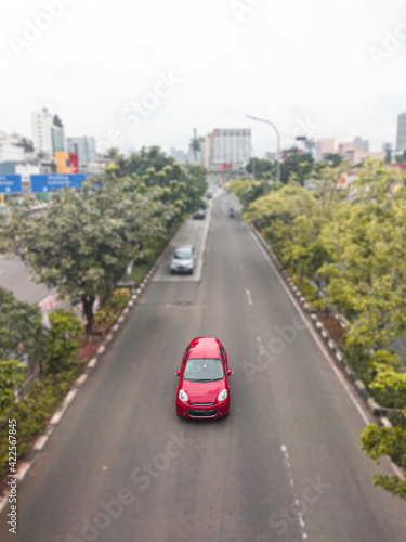 A beautiful red car crosses the middle of Jakarta Indonesia s road  on the left and right of the road  there are shady trees  making the road more comfortable for vehicles to pass.