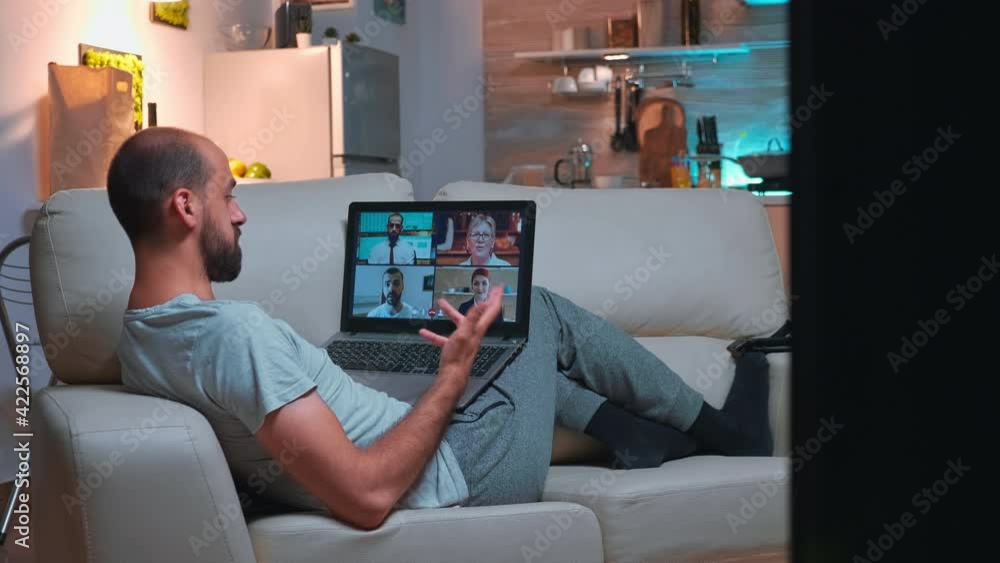 Business man in pajams lying on sofa while chatting with friends during videocall meeting using laptop computer. Caucasian male using modern technology wireless late at night in kitchen Business man in pajams lying on sofa while chatting with friends during videocall meeting using laptop computer. Caucasian male using modern technology wireless late at night in kitchen