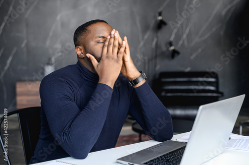 Exhausted African-American male employee covered face with a palms, tired black businessman sits at workplace with a laptop, his eyes closed, overwork concept. Frustrated man has problems with project photo