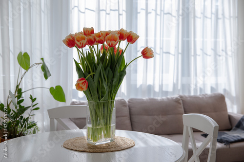 A bright bouquet of tulips in the interior of a light kitchen. Vase with tulips on the table. Spring flowers. Fresh orange buds. Cozy kitchen. Flowers on the background of the window