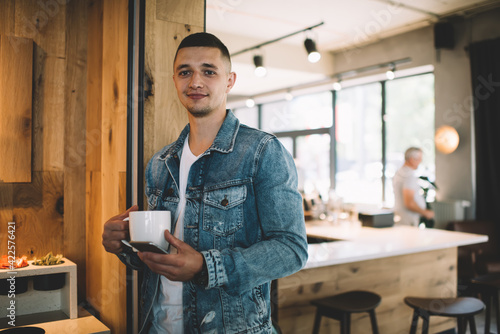 Smiling young man with cup of coffee and smartphone in cafe
