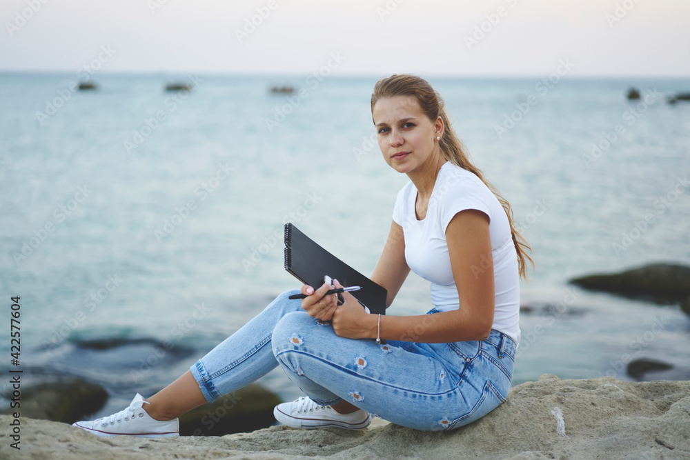 Portrait of Caucasian female artist with education sketchbook resting at rock at seashore and looking at camera during leisure time for learning, talented student with notepad posing on weekend