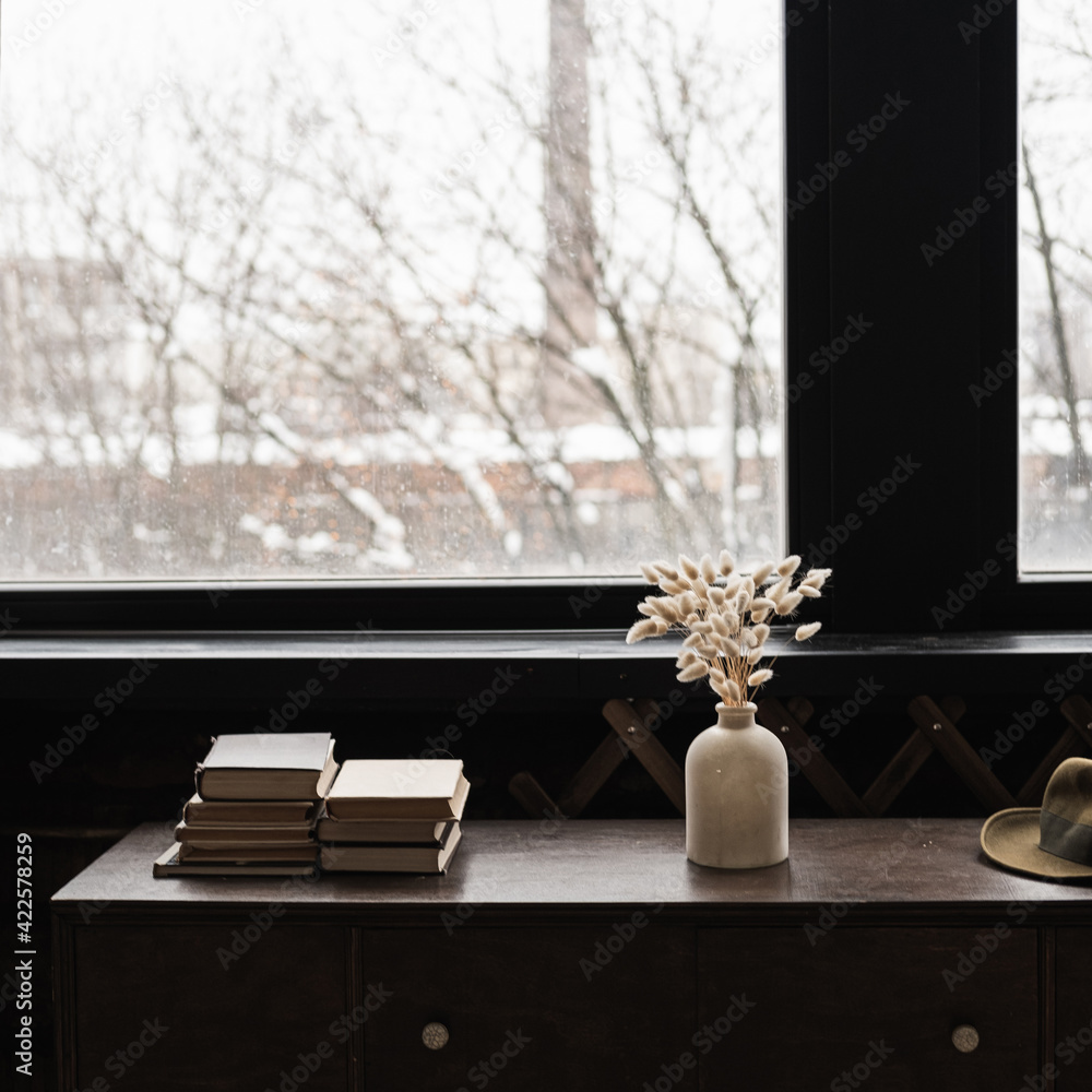 Fototapeta premium Rabbit bunny tail grass bouquet, books stack on solid wooden table against window. Aesthetic dark home interior design concept. Beautiful floral foliage composition.