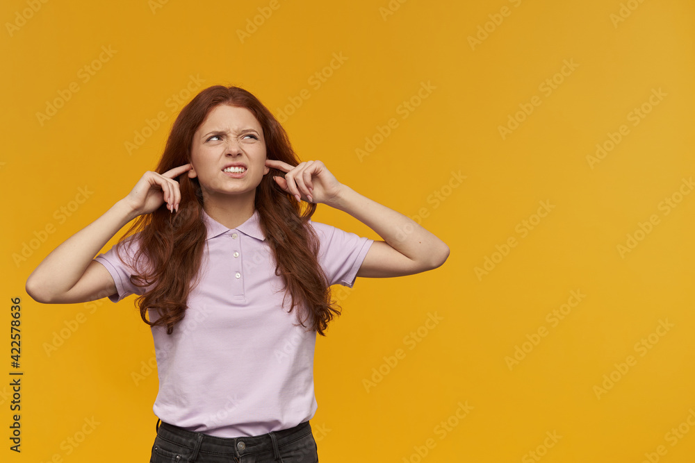 Portrait of annoyed, frowning lady with long ginger hair. Wearing pink t-shirt. Close ears with fingers, irritated with noise. Watching to the right at copy space, isolated over orange background Portrait of annoyed, frowning lady with long ginger hair. Wearing pink t-shirt. Close ears with fingers, irritated with noise. Watching to the right at copy space, isolated over orange background