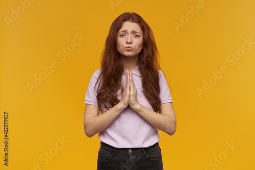 Portrait of regretting, sad lady with long ginger hair. Wearing pink t-shirt. Emotion concept. Keeps palms in prayer, plead for something. Watching at the camera, isolated over orange background © timtimphoto