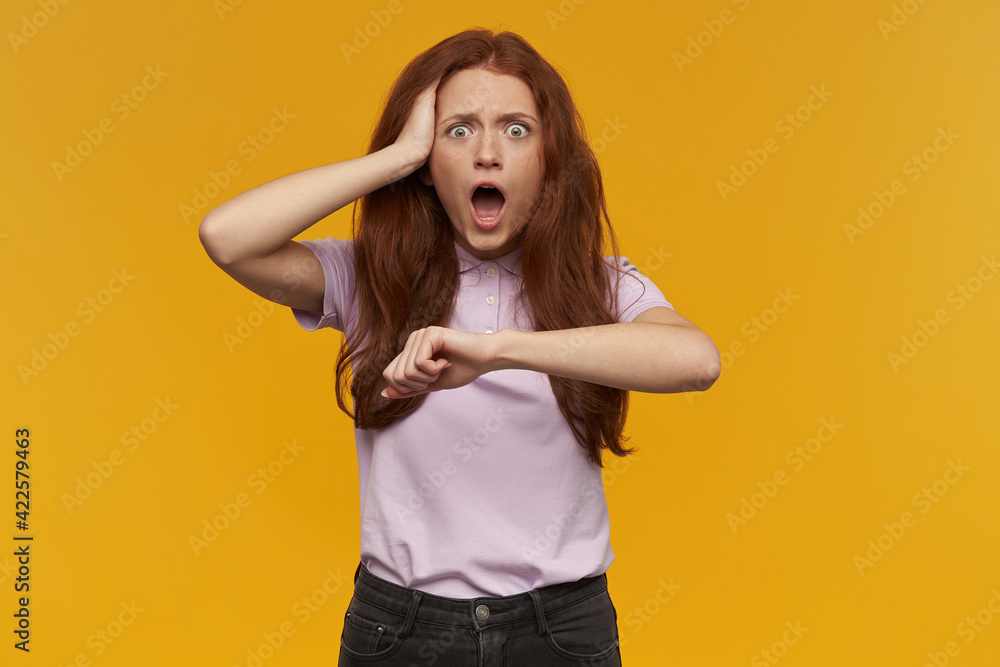 Stressed, shocked woman with long ginger hair. Wearing pink t-shirt. Forgot about time. Touching her head and imitate a wrist watch. Watching at the camera, isolated over orange background Stressed, shocked woman with long ginger hair. Wearing pink t-shirt. Forgot about time. Touching her head and imitate a wrist watch. Watching at the camera, isolated over orange background