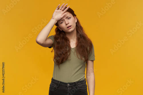 Portrait of exhausted, tired lady with long ginger hair. Wearing green t-shirt. People and emotion concept. Touching her forehead, feels sick. Watching at the camera, isolated over orange background