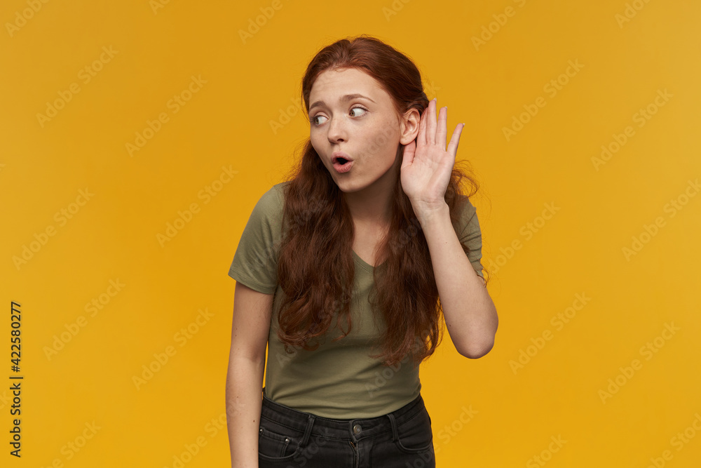 Portrait of curious, beautiful lady with long ginger hair. Wearing green t-shirt. People and emotion concept. Keeps palm next to ear, eavesdrop conversation. Stand isolated over orange background Portrait of curious, beautiful lady with long ginger hair. Wearing green t-shirt. People and emotion concept. Keeps palm next to ear, eavesdrop conversation. Stand isolated over orange background