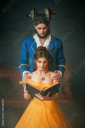 Fantasy man enchanted prince with ram's horns on head, hugging a beautiful woman by shoulders. Girl in yellow medieval historical dress holds reads vintage book. Gothic dark room. Guy beast monster photo