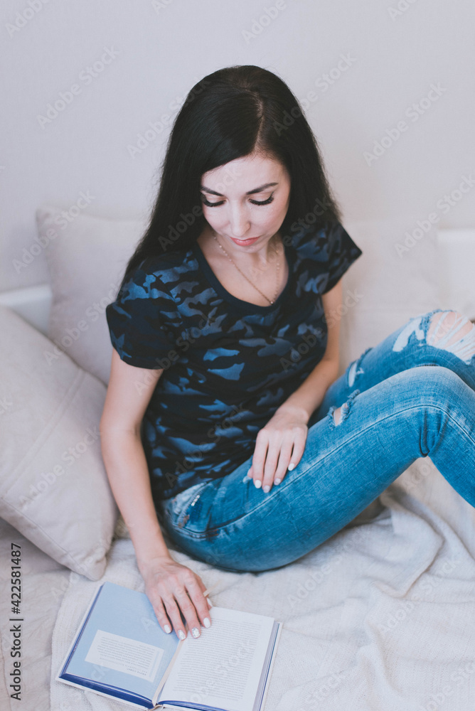 brunette girl in a t-shirt and blue jeans reads a book on the bed brunette girl in a t-shirt and blue jeans reads a book on the bed
