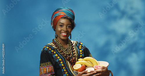 Portrait shot of young cheerful smiled pretty African American woman demonstrating plate with tropical fruit and looking at camera. Joyful beautiful girl showing fruits and smiling happily. photo