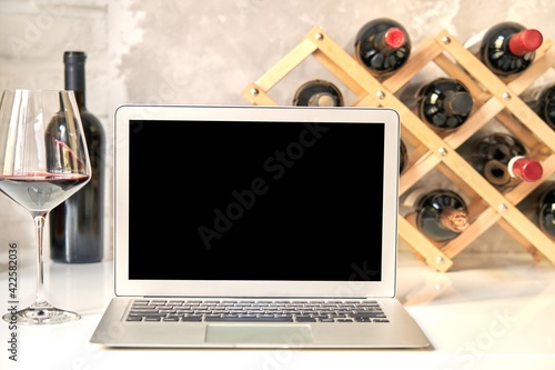 Empty blank screen laptop computer on table with wine bottle wine rack holder in the background. Buying wines online, home delivery concepts.