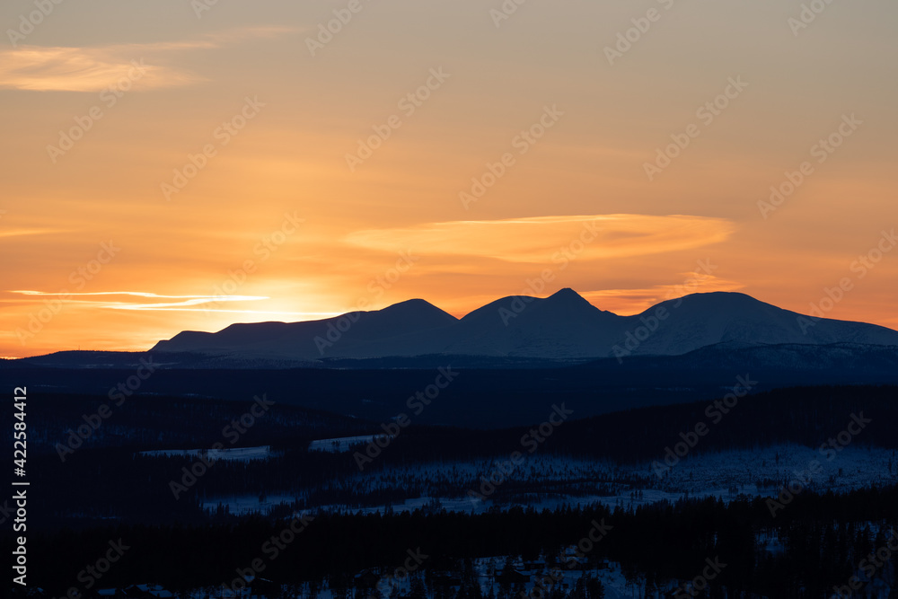 Obraz premium Sunset behind Norwegian mountains in winter. Orange sky and snow covered woodland. Shot in Sweden, Scandinavia
