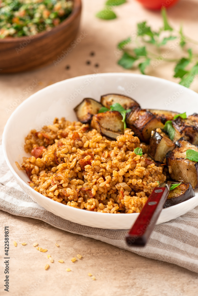 Bulgur pilaf (pilavi) with tomatoes and fried eggplants in a white plate on a light table close-up. Turkish cuisine, vegan food. Bulgur pilaf (pilavi) with tomatoes and fried eggplants in a white plate on a light table close-up. Turkish cuisine, vegan food.