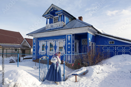 Russian girl in Pavlovo Posad shawl. Wooden rural house with carved windows in Purekh village, Nizhny Novgorod region, Russia. Snow winter. Russian folk style in architecture, architectural fashion photo